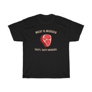 Op de afbeelding: Zwart t-shirt met de tekst "MEAT IS MURDER" boven een cartoon steak illustratie. Onder de illustratie staat de tekst "TASTY, TASTY MURDER."