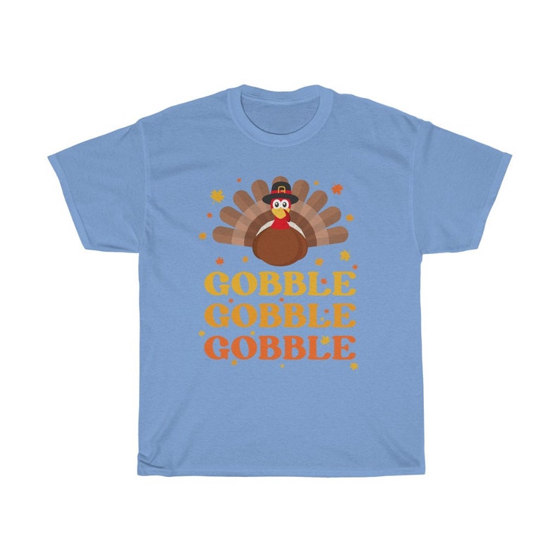 May include: Light blue t-shirt featuring a cartoon turkey wearing a pilgrim hat, with the words "GOBBLE GOBBLE GOBBLE" in orange and yellow. The design includes autumn leaves. A festive graphic tee for Thanksgiving.