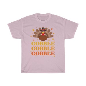 May include: A light pink t-shirt featuring a cartoon turkey wearing a pilgrim hat, surrounded by autumn leaves. The words "GOBBLE GOBBLE GOBBLE" are stacked in orange and yellow. A festive design for Thanksgiving.