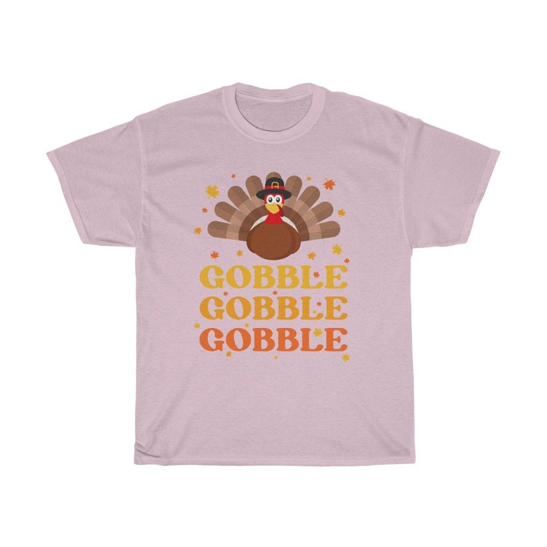 May include: A light pink t-shirt featuring a cartoon turkey wearing a pilgrim hat, surrounded by autumn leaves. The words "GOBBLE GOBBLE GOBBLE" are stacked in orange and yellow. A festive design for Thanksgiving.