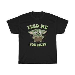 May include: Black t-shirt featuring a cartoon character with large eyes and green skin, wearing a brown coat. The text "FEED ME YOU MUST" is printed above and below the character in a retro font.