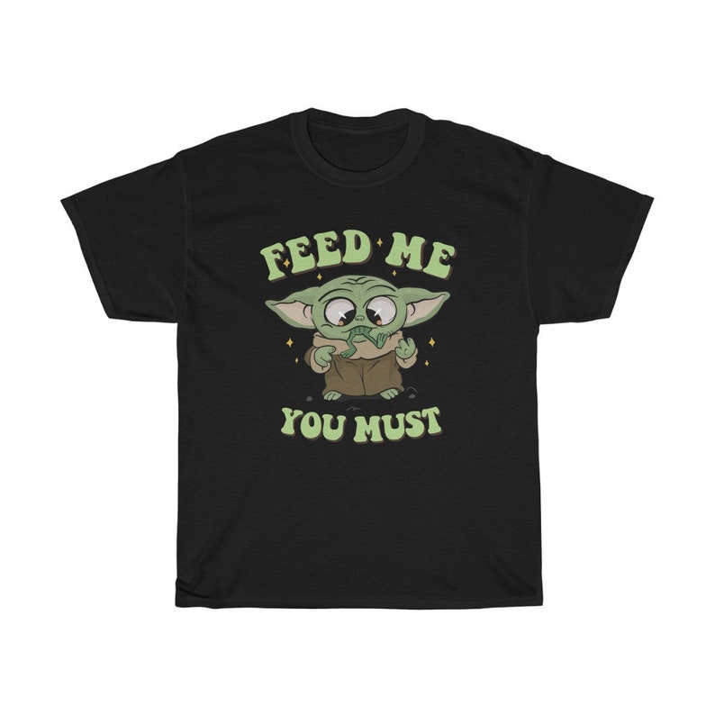 May include: Black t-shirt featuring a cartoon character with large eyes and green skin, wearing a brown coat. The text "FEED ME YOU MUST" is printed above and below the character in a retro font.