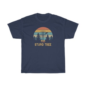 Pode incluir: T-shirt azul marinho com um gr&aacute;fico retr&ocirc; de uma cesta de disc golf, um disco e &aacute;rvores contra um p&ocirc;r do sol. O texto "STUPID TREE" est&aacute; abaixo do gr&aacute;fico. Uma camiseta casual para os entusiastas do disc golf.