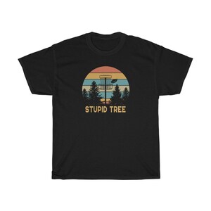 Pode incluir: Camiseta preta com um design retr&ocirc; com uma cesta de disc golf, um disco voador e pinheiros contra um p&ocirc;r do sol. O texto "STUPID TREE" est&aacute; abaixo.