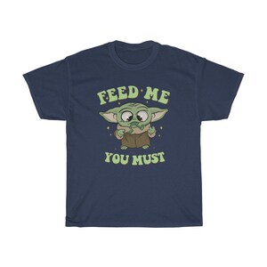 May include: Navy blue t-shirt featuring a cartoon character with large eyes and green skin. The character is holding a green object and the text "FEED ME YOU MUST" is above and below the character in green lettering.