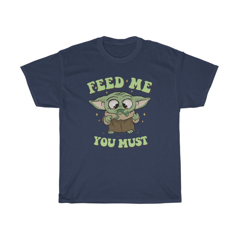 May include: Navy blue t-shirt featuring a cartoon character with large eyes and green skin. The character is holding a green object and the text "FEED ME YOU MUST" is above and below the character in green lettering.