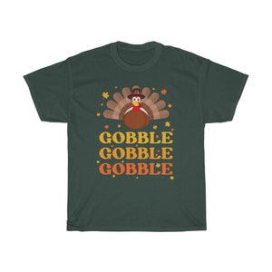 May include: A dark green t-shirt featuring a cartoon turkey wearing a pilgrim hat, with the words "GOBBLE GOBBLE GOBBLE" in orange and yellow. The design includes fall leaves. A festive shirt for Thanksgiving.