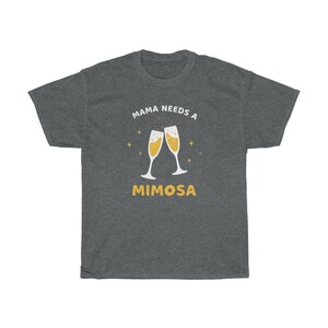 May include: Dark gray t-shirt with the text "MAMA NEEDS A MIMOSA" in white and yellow. The design features two champagne flutes with bubbly liquid and star accents. A casual, comfortable tee.