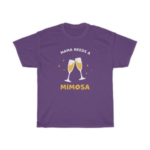 May include: A purple t-shirt with the text "MAMA NEEDS A MIMOSA" in white and yellow. The design features two champagne flutes clinking together, filled with a bubbly drink, and surrounded by small starbursts.