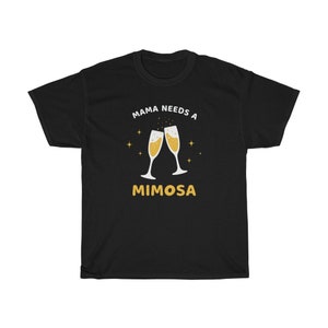 May include: Black t-shirt with the text "MAMA NEEDS A MIMOSA" in white and gold. The design features two champagne flutes with bubbly drinks and gold stars. A fun and casual tee for a relaxed vibe.