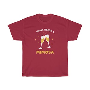 May include: A maroon t-shirt with the text "MAMA NEEDS A MIMOSA" in white and gold. The design features two champagne flutes clinking together, with gold star accents. A casual, comfortable tee.
