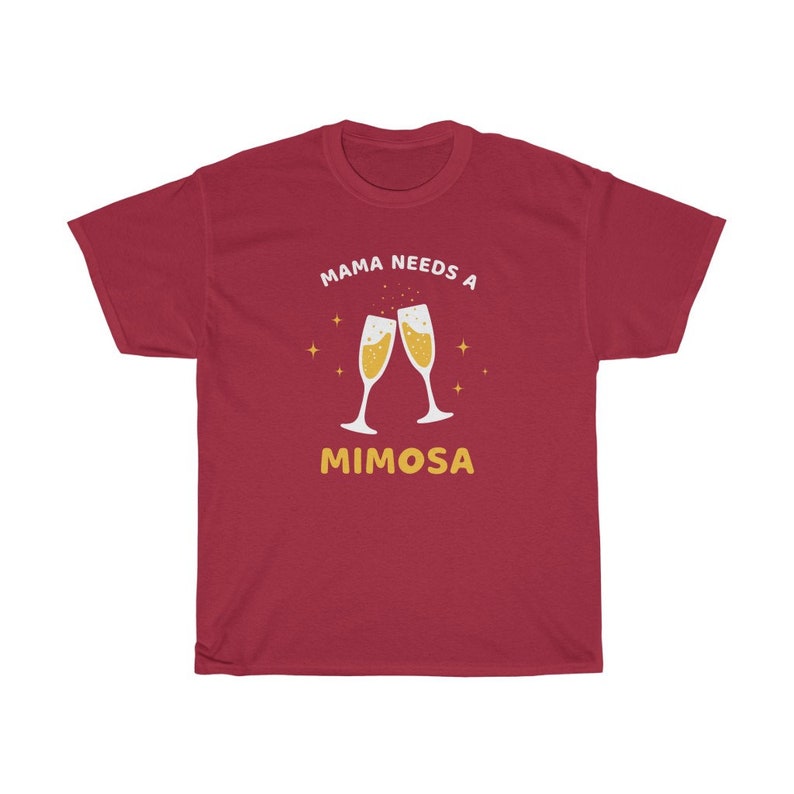 May include: A maroon t-shirt with the text "MAMA NEEDS A MIMOSA" in white and gold. The design features two champagne flutes clinking together, with gold star accents. A casual, comfortable tee.