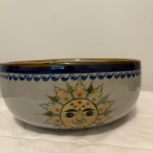 May include: A ceramic bowl with a light gray base color featuring a hand-painted sun face design in yellow, white, and black. The rim is decorated with a dark blue wavy line pattern, and the interior edge shows a brown glazed finish.