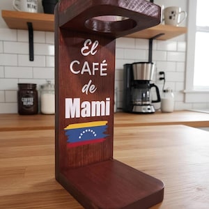 May include: A dark brown wooden pour-over coffee stand with a circular top opening. The front panel features the white text "El CAFÉ de Mami" above a painted graphic of the Venezuelan flag with yellow, blue, and red horizontal stripes and stars.
