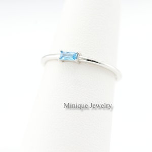 May include: A silver ring with a rectangular, light blue gemstone. The ring is delicate and minimalist, with the text "Minique Jewelry" in a light gray font.