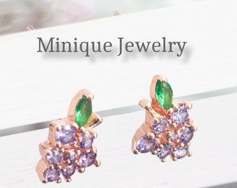 Könnte beinhalten: Ein Paar goldfarbene Ohrringe in Form von Trauben mit lila und grünen Zirkonia-Steinen. Die Ohrringe befinden sich auf einer weißen Oberfläche mit dem Text "Minique Jewelry" darüber.
