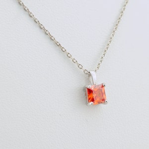 May include: A silver chain necklace with a square-shaped pendant featuring a bright orange gemstone.