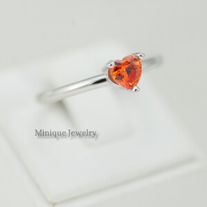 May include: A silver ring with a heart-shaped orange gemstone. The ring is on a white background. Minique Jewelry.