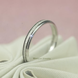 May include: A silver ring with a textured, glittery band. The ring is set against a soft pink background and rests on a white cloth. The ring is from Minique Jewelry.
