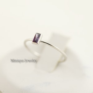 May include: A delicate silver ring with a small, rectangular purple gemstone. The ring is simple and elegant, perfect for everyday wear.