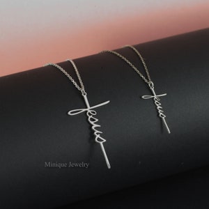 May include: Two silver necklaces with a cross pendant featuring the word "Jesus" in cursive script. The necklaces are on a black surface.