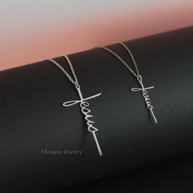 May include: Two silver necklaces with a cross pendant featuring the word "Jesus" in cursive script. The necklaces are on a black surface.
