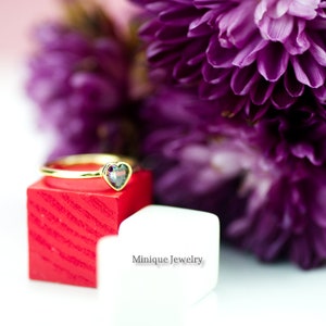 May include: A gold ring with a heart-shaped iridescent gemstone sits on a red and white display box. The ring is surrounded by purple flowers.