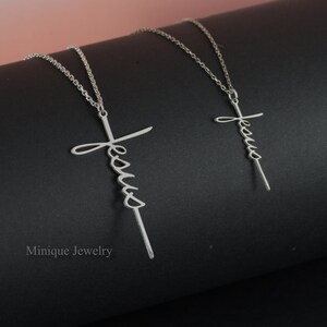 May include: Two silver necklaces with a cross pendant featuring the word "Jesus" written in cursive. The necklaces are on a black surface.