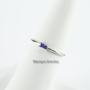 May include: A delicate silver ring with a small, rectangular purple gemstone. The ring is displayed on a white stand. "Minique Jewelry" is written below the ring.