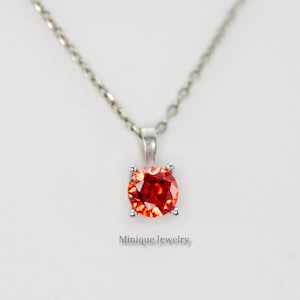 May include: A silver chain necklace with a red gemstone pendant. The pendant is set in a silver bezel and has a round shape. The text "Minique Jewelry" is visible below the pendant.