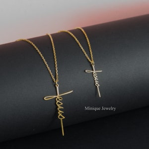 May include: Two gold necklaces with a cross pendant featuring the word "Jesus" written in cursive. The necklaces are on a black surface. Minique Jewelry.