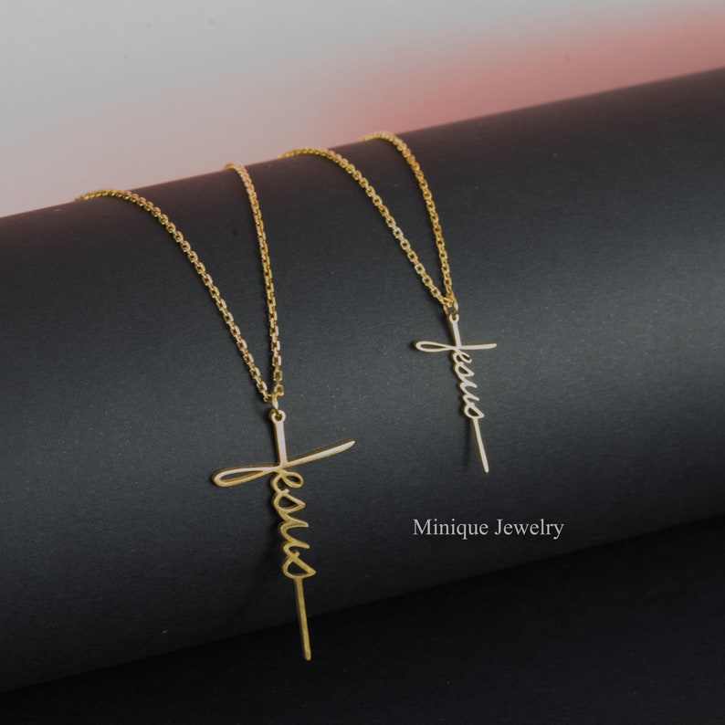 May include: Two gold necklaces with a cross pendant featuring the word "Jesus" written in cursive. The necklaces are on a black surface. Minique Jewelry.