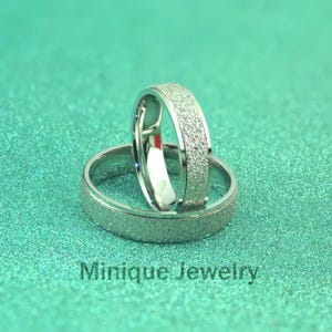 May include: Two silver rings with a textured band and a smooth interior are displayed on a teal, glittery surface. The rings are slightly angled, showcasing their design. The text "Minique Jewelry" is visible at the bottom.