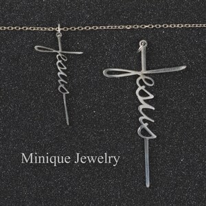 May include: Two silver necklaces with a cross pendant featuring the word "Jesus" in a cursive script. The necklaces are on a black background.