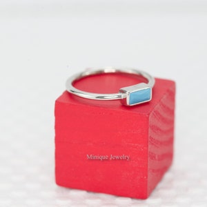 May include: A silver ring with a light blue rectangular stone set on a red wooden block. The block has the text "Minique Jewelry" printed on it.