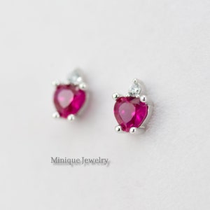 May include: A pair of silver heart-shaped earrings with a small clear stone on top of each heart. The hearts are filled with a bright pink stone.