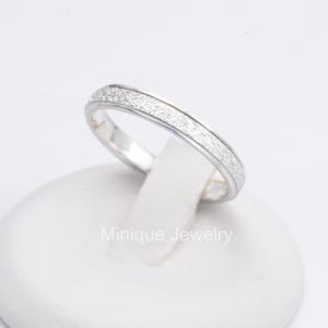 May include: A silver ring with a textured band of small, sparkling stones. The ring is displayed on a white stand against a white background. The text "Minique Jewelry" is visible on the stand.