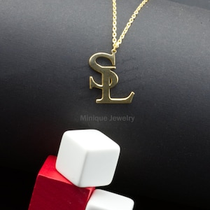 May include: Gold-coloured pendant necklace with the letters "SL" in a stylized font. The necklace hangs on a delicate chain against a dark background. The brand name "Minique Jewelry" is printed below the pendant. White and red cubes are in the foreground.