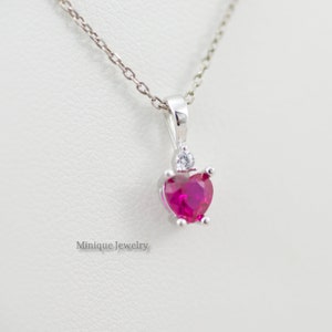 May include: A silver necklace with a heart-shaped pendant featuring a pink gemstone and a smaller clear gemstone. The pendant is set in a silver setting with a delicate chain.