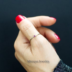May include: A delicate silver ring with a small, round, ruby-colored gemstone. The ring is displayed on a finger with red nail polish, against a dark background. The ring is a minimalist design, perfect for everyday wear. The text "Minique Jewelry" is visible.