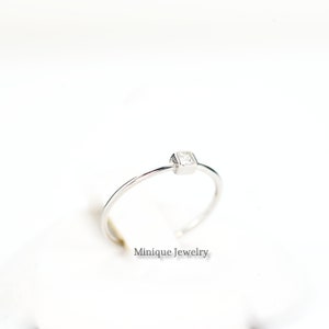 May include: A minimalist silver ring with a square, clear gemstone. The ring has a thin band and a simple, modern design. The text "Minique Jewelry" is visible.