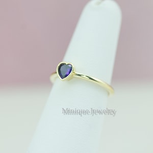 May include: A gold ring with a small purple heart-shaped gemstone. The ring is on a white display stand.