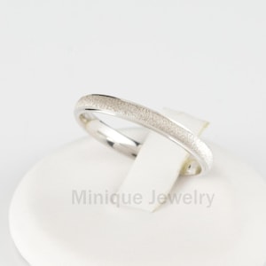 May include: A thin silver band with a textured finish. The ring is displayed on a white stand with the text "Minique Jewelry".
