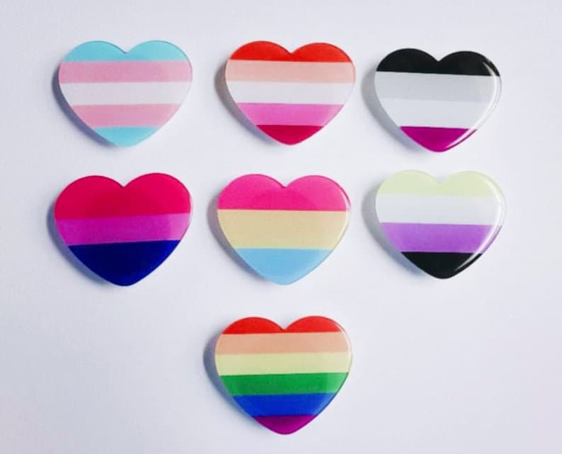 K&ouml;nnte beinhalten: Sieben herzf&ouml;rmige Pins mit verschiedenen Designs von Pride-Flaggen. Die Flaggen umfassen die Transgender-, Lesben-, Bi-, Pan-, Genderqueer-, Asexual- und Regenbogen-Pride-Flaggen.