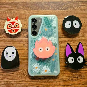 May include: A light blue phone case with a floral design, surrounded by character-themed phone grips. The grips include a red and white mask, a black soot sprite, a black cat with purple ears, a pink flame, and a black and white face.