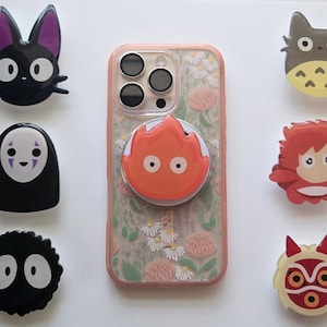 May include: A smartphone in a floral patterned case with a circular orange flame character grip attached to the back. Surrounding the phone are six circular character-themed grips featuring a black cat, a white ghost, a black soot sprite, a grey creature, a red-haired figure, and a red-masked character.