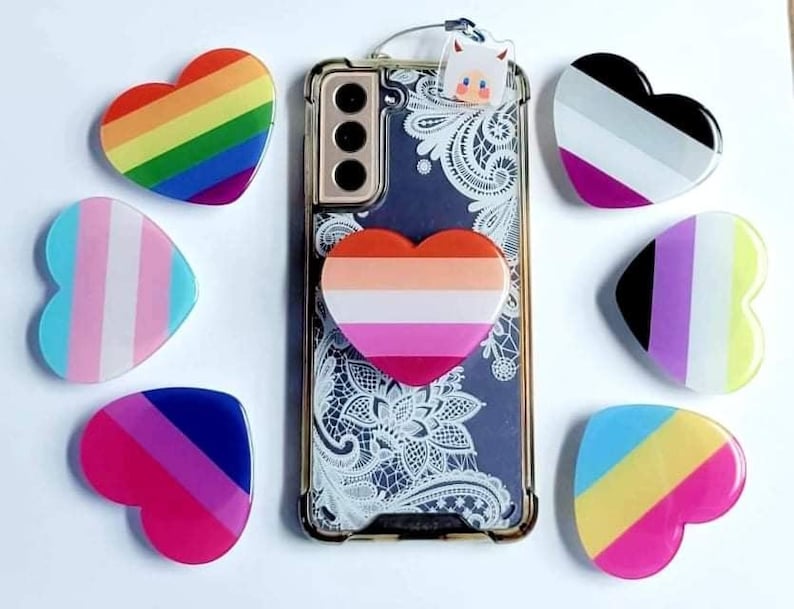 K&ouml;nnte beinhalten: Eine Handyh&uuml;lle mit verschiedenen herzf&ouml;rmigen Handygriffen. Die Griffe sind in verschiedenen Farben und Mustern, darunter Regenbogen, Transgender, Bisexuell, Pansexuell und Asexuell-Pride-Flaggen.