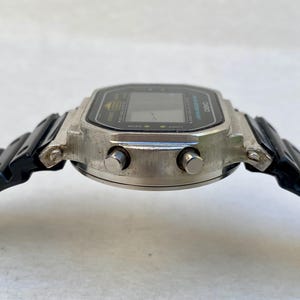 May include: A vintage digital watch with a silver case and a black strap. The watch face is black with a digital display and buttons. The strap is black plastic. The watch has a classic retro design, reminiscent of the 1980s.