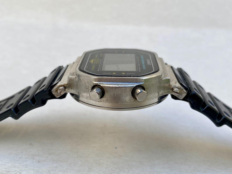 May include: A vintage digital watch with a silver case and a black strap. The watch face is black with a digital display and buttons. The strap is black plastic. The watch has a classic retro design, reminiscent of the 1980s.