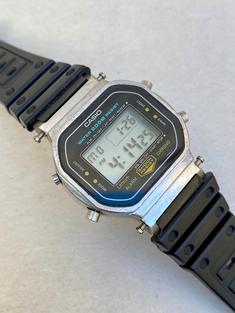 May include: A Casio digital watch with a black and silver rectangular face and a black strap. The watch displays the time and date. The face has the text "CASIO WATER 200M RESIST".